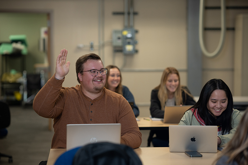 A smiling student in a brown shirt raises a hand in class
