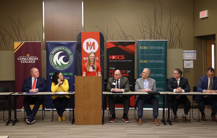 Student speaker Carley Berg speaks at the podium with (seated, left to right) Tri-College Provost Tim Flakoll, M State President Carrie Brimhall, MSUM President Timothy Downs, NDSCS President Rod Flanigan, Concordia College President Colin Irvine and NDSU President David Cook.