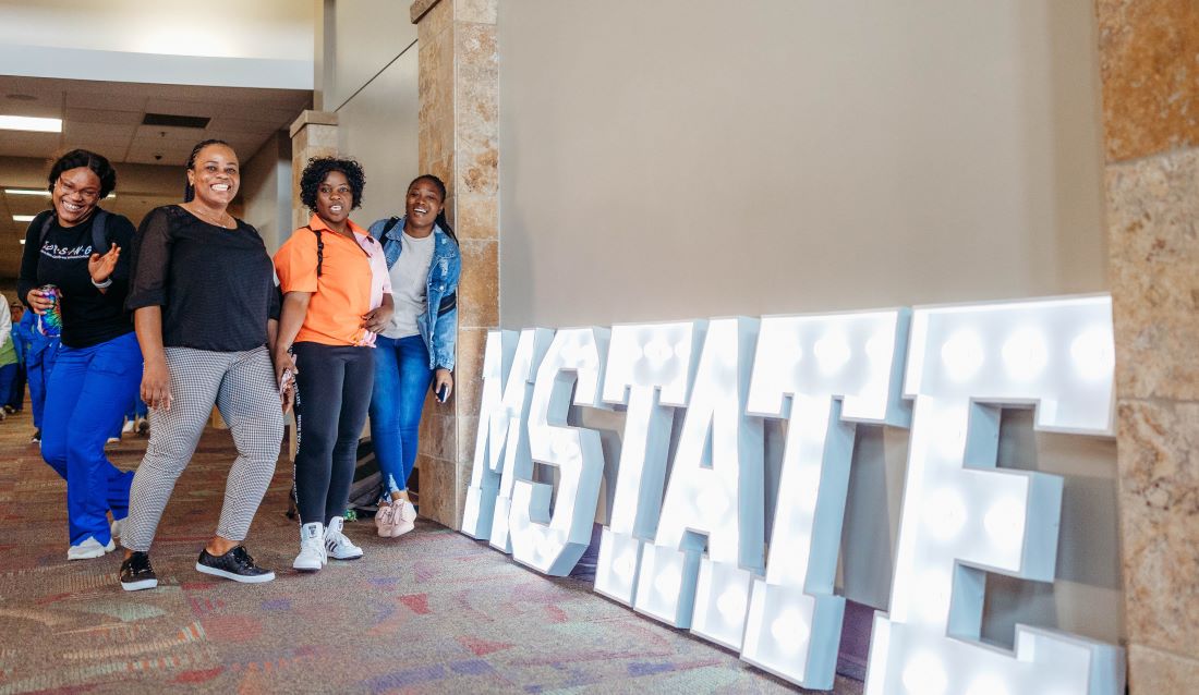 Four M State students laugh and pose in front of a large light-up M State sign