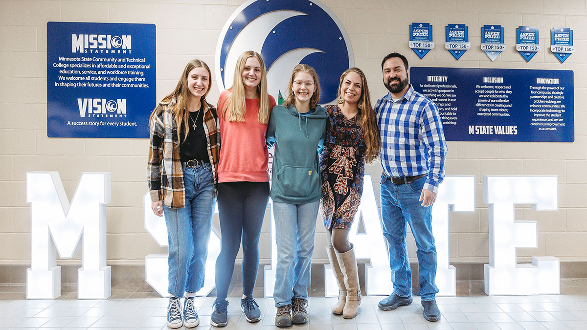 A group of students stands in front of an M State sign at the Detroit Lakes campus