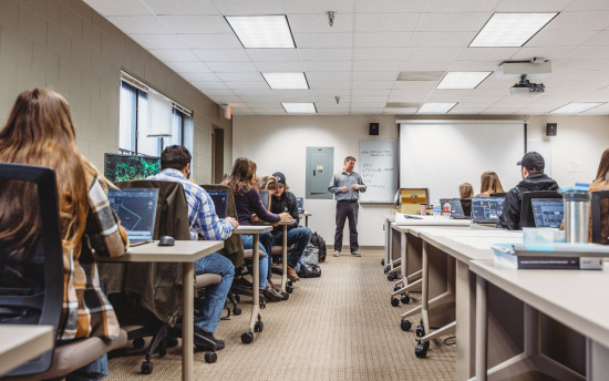 A classroom full of students with the instructor at the front of the room drawing their attention