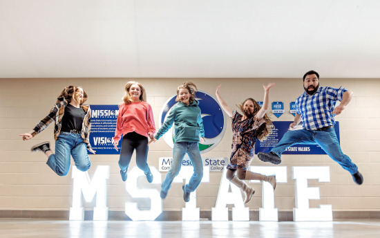 Five students jumping for joy over a large, light-up M State sign