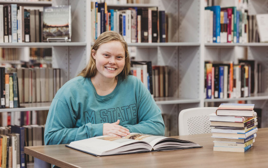 A student in a blue M State sweatshirt in the library smiles