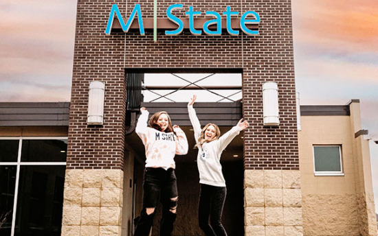 Two students jump in front of an M State entrance