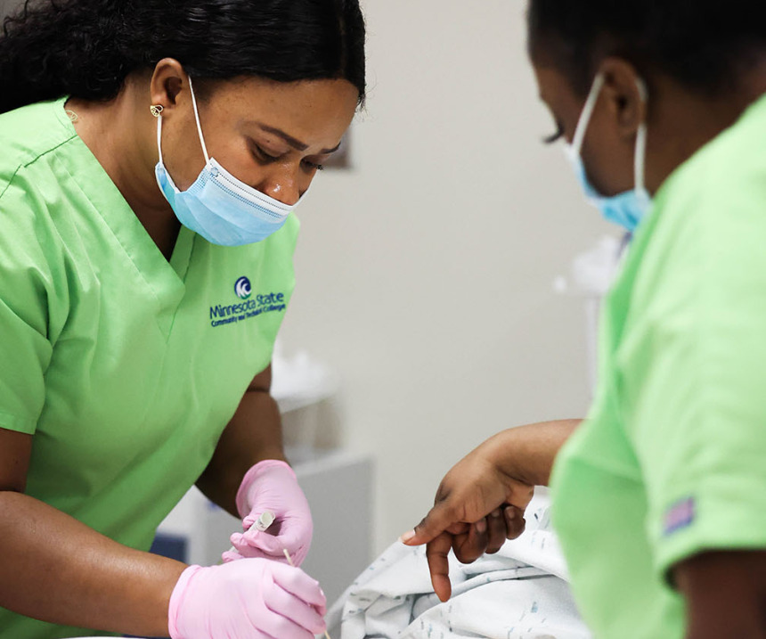 Two students in green scrubs work on a mannequin in class